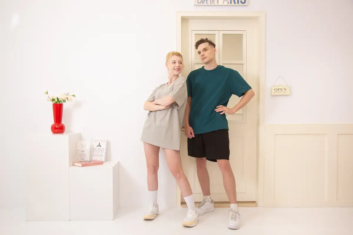 ARRIVE GUIDE AG18000頂級精梳棉T恤 一對男女模特兒穿著 AG18000 圓領精梳棉 T 恤,展示燕麥色長版穿搭與孔雀綠休閒穿搭,背景為簡約白色室內場景。