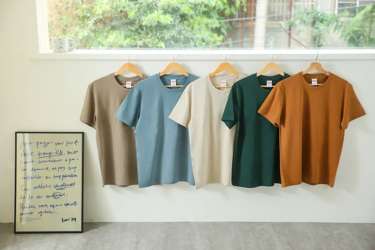 ARRIVE GUIDE AG18000頂級精梳棉T恤 五件不同顏色的 AG18000 精梳棉 T 恤掛在白牆窗邊,顏色包含摩卡棕、霧灰藍、燕麥色、深綠色與焦糖色,展現豐富的莫蘭迪色系選擇。