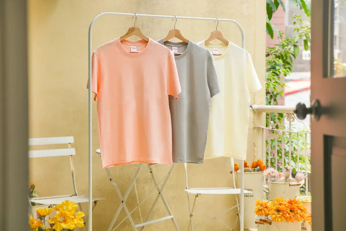 ARRIVE GUIDE AG18000頂級精梳棉T恤 三件 AG18000 精梳棉 T 恤(粉色、灰色、奶白色)吊掛在戶外陽台的白鐵衣架上,陽光透過植物灑落,呈現清新自然的居家氛圍。